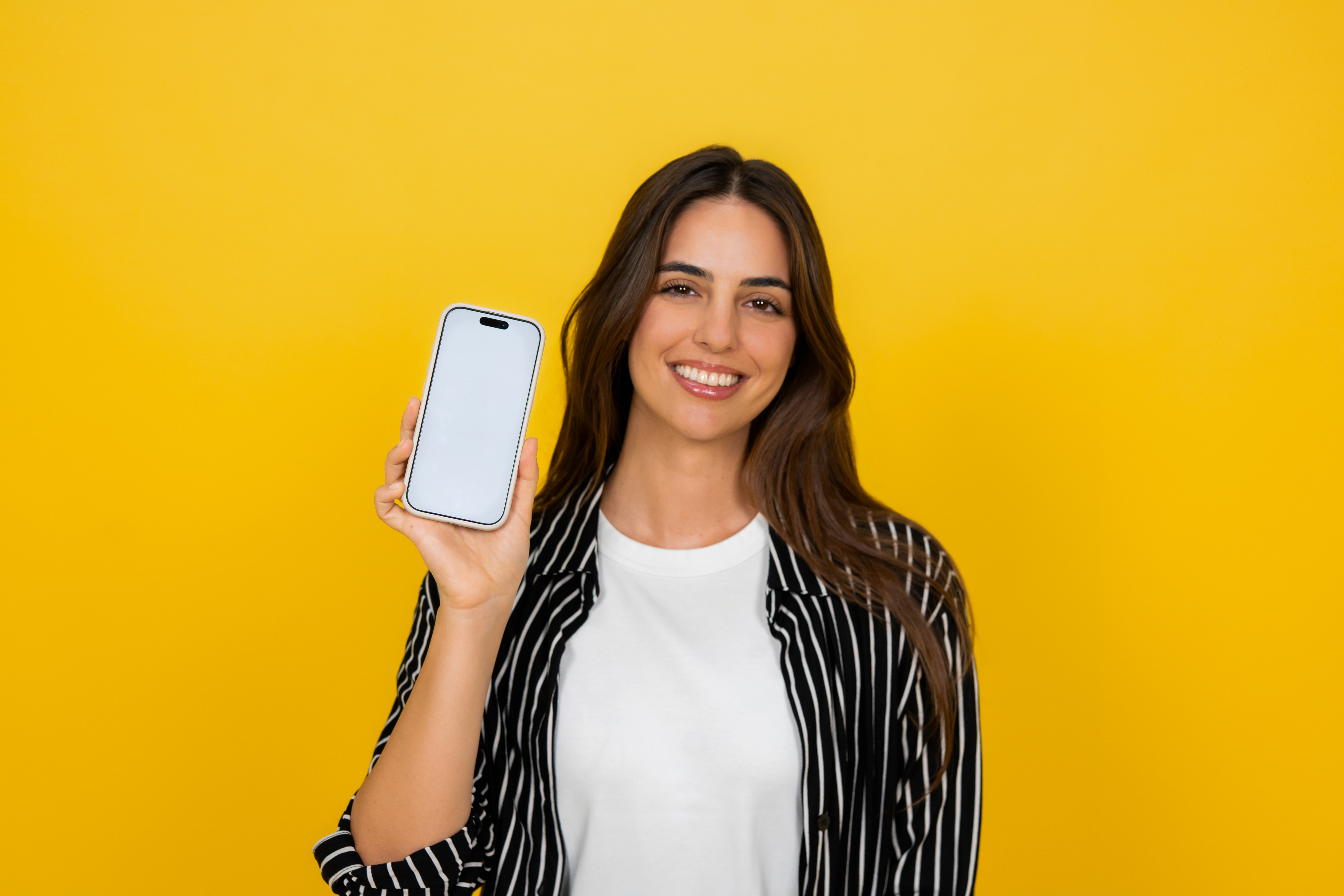 Young adult woman holding a modern smartphone with a blank white screen, presenting it to the viewer while smiling brightly on a vibrant yellow studio background